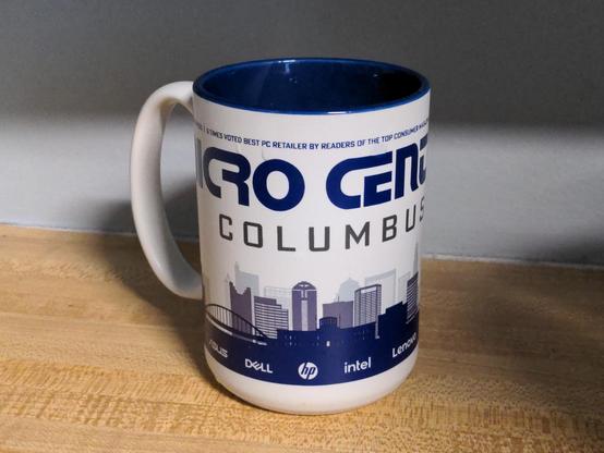 Coffee mug on a counter. This side reads "Micro Center Columbus", featuring a silhouette of a city skyline and logos for Dell, HP, Intel, and Lenovo.