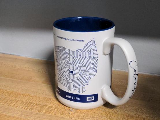 Coffee mug on a counter. This side reads featuring a drawing of the State of Ohio made to look like a circuit board, with a chip where Columbus is located. Also there are logos of Samsung and Western Digital, and the handle has the word Columbus written in cursive.