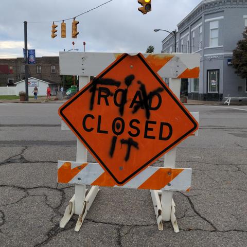 Orange diamond shaped traffic sign mounted on a wooden frame, sitting in the middle of a town intersection. Black letters on the sign read "Road closed", but someone has spray painted over it, in black paint,  the words 'Fix it".