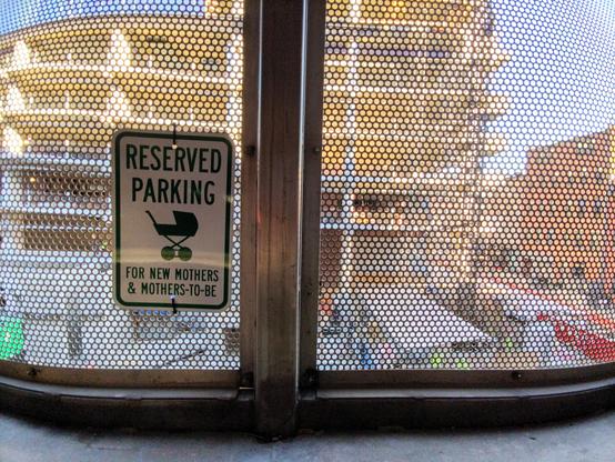 Metal sign with an icon of a baby stroller reading "Reserved parking for new mothers and mothers to be". The sign hangs on a metal grilled wall or fence that can be partially seen through. Inside the wall it's fairly dark, but outside it's sunny and bright. The view is taken up by construction of a building at least 6 stories tall, next to a city street and a 5 story building. The building being constructed looks to be a car parking garage, everywhere open to the outside rather than walled off.…