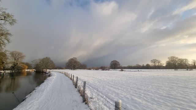 Photo of little river with snowy footpath and fields. Trees in the distance. Low morning sun with clouds.