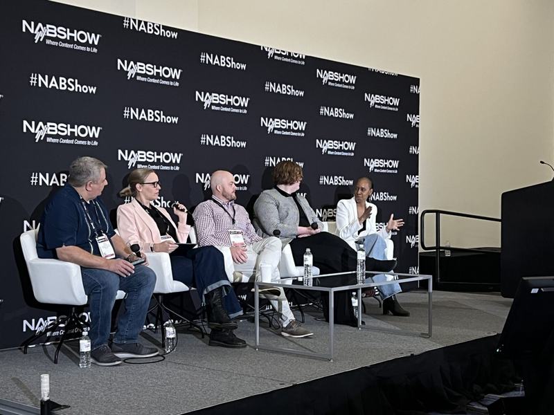 One moderator and four panelists sit on stage at the NAB Show in front of a black backdrop with repeated “#NABShow” and “Where Content Comes to Life” logos. The panelists are seated in white chairs with microphones and water bottles in front of them. 