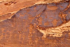 Ancient Native American petroglyphs etched into a red rock wall near Moab, Utah, showing horned figures, animals, and symbolic shapes.