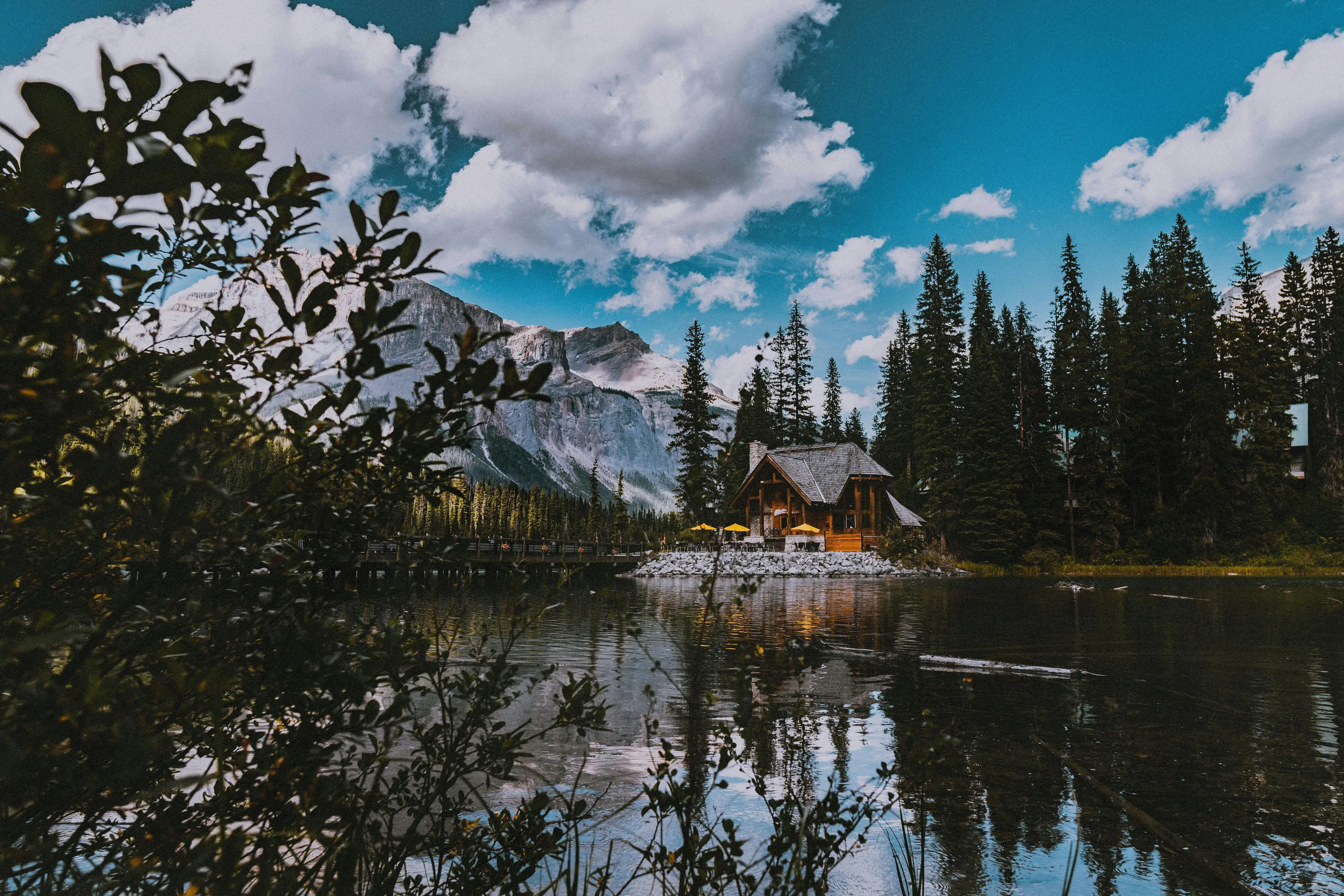 A serene cabin is nestled amongst pines, surrounded by a calm lake with snowy mountains behind it.