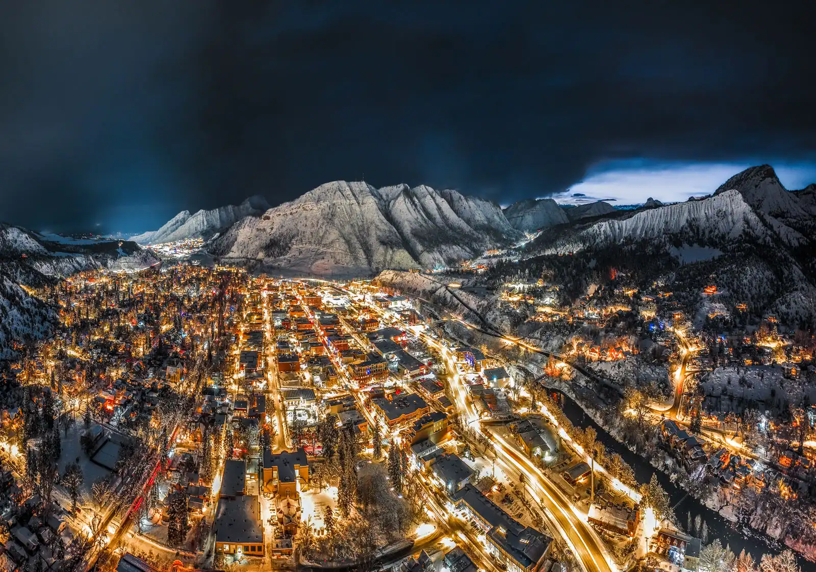 Aerial night view of Durango, Colorado, with illuminated streets and snow-covered San Juan Mountains under a stormy sky.