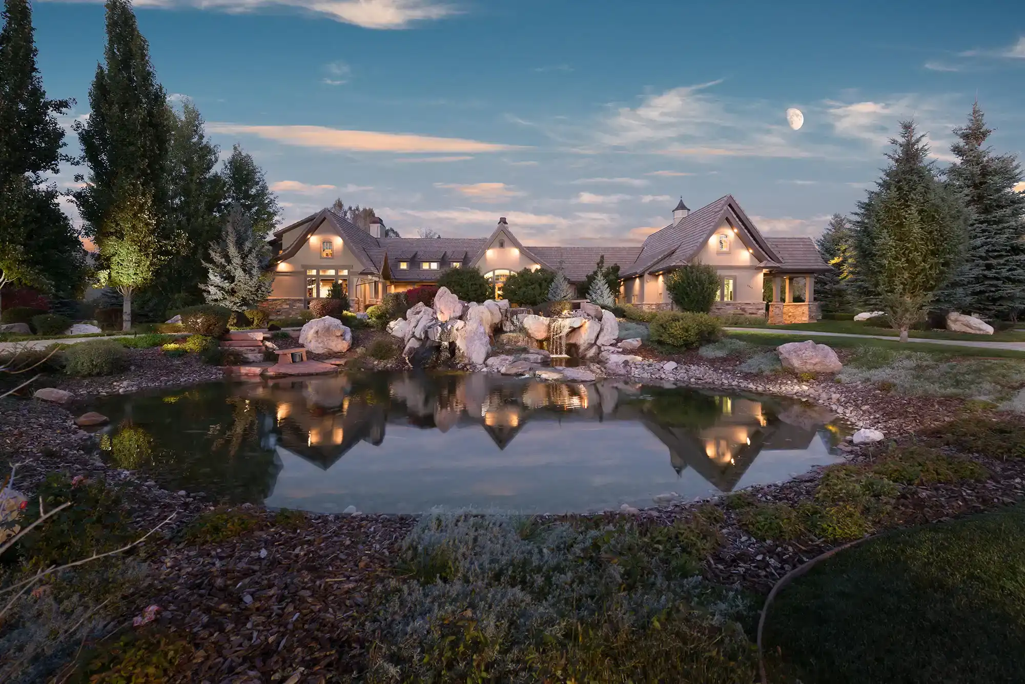 A luxury home under the moon at twilight with a lit pond in the front yard.