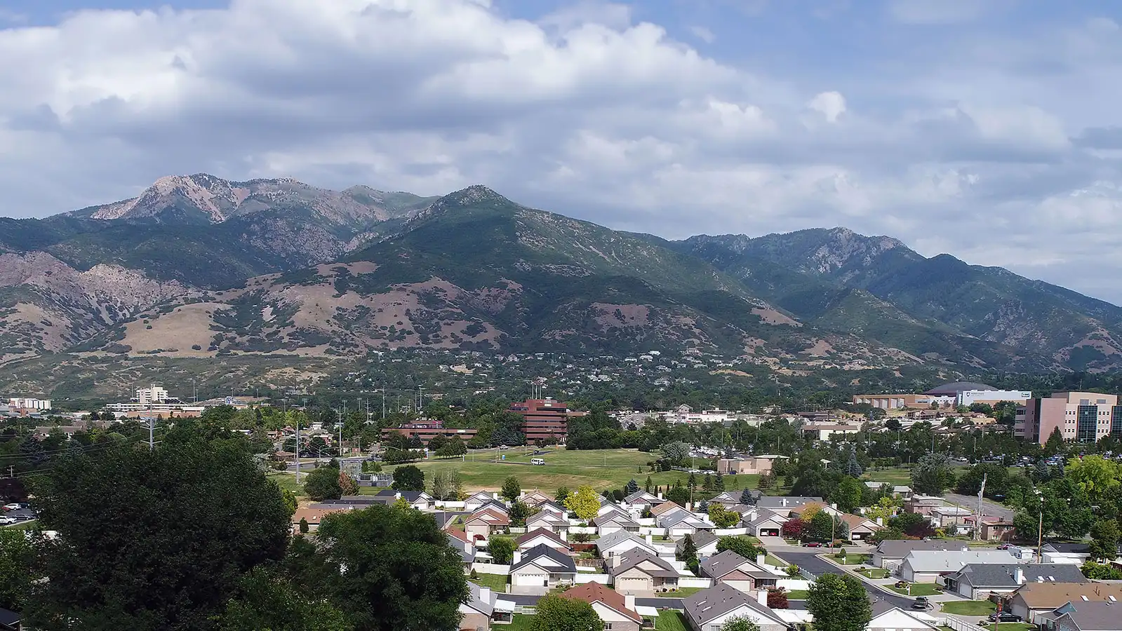 Panoramic view of South Ogden, Utah, with residential neighborhoods in the foreground and the verdant Wasatch Mountains rising majestically in the background under a cloudy sky.