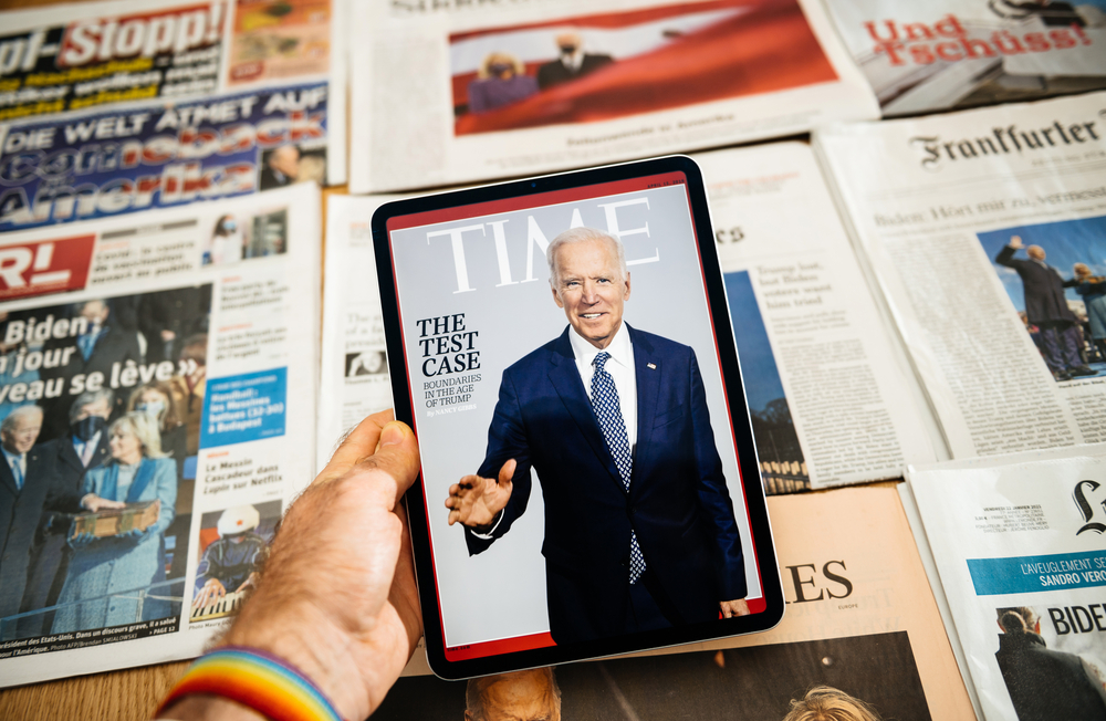 man holding digital tablet with time magazine cover featuring joe biden and stack with international newspapers