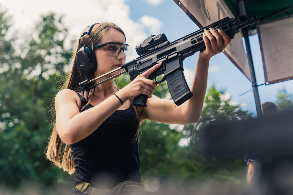 strong powerful confident long haired caucasian woman in a black tank top holding big black rifle with loupe preparing for a shooting competition. medium outdoor shot.