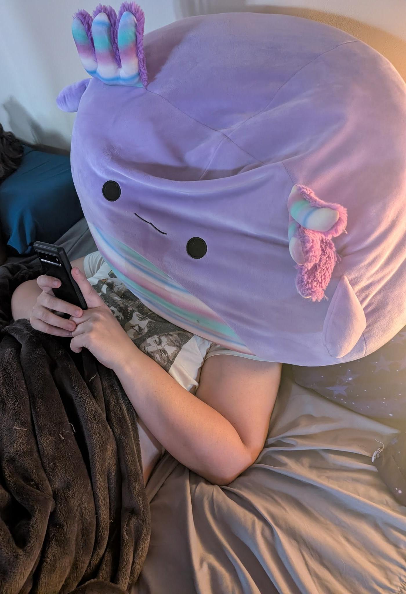 white girl in a bed on her phone, except a giant round purple stuffed animal covers her face, and it looks like the stuffed animal is on the phone.