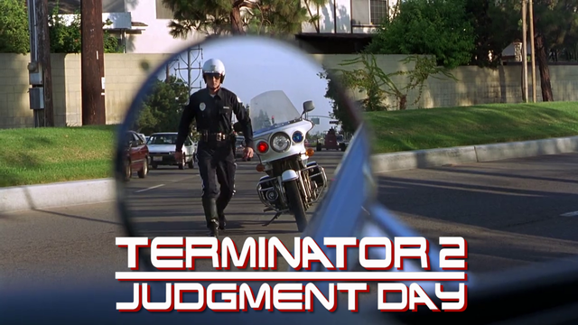 A screenshot from a movie, labelled Terminator 2: Judgment Day. It shows side mirror of a blue car (some would say underpants blue). Mirrored is Robert Patrick from T2 dressed as a cop walking from his motorcycle towards the blue car car.