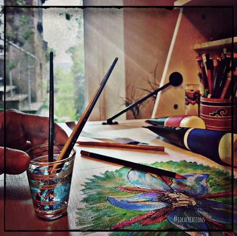 Une table de travail, éclairée par une porte fenêtre donnant sur un jardin. Je tiens un petit verre prévu pour le thé à la menthe. Il sert pour des pinceaux. Ils ont teinté l'eau en bleu. Des crayons sont éparpillés. Un dessin de fleur attend d'être terminé.