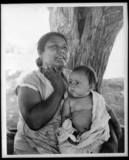 The image is a black and white photograph featuring an older woman seated outdoors with her arms around a baby she's holding. They appear to be sitting against the trunk of a large tree, which provides partial shade over them. The woman has dark skin and wears earrings; she gazes upward with a contemplative expression on her face as if lost in thought or reflection. She is wearing what appears to be a sleeveless blouse with a checkered pattern around the shoulders.

The baby looks slightly pensive but also attentive, looking off into space away from the camera's perspective. The child has dark hair and seems relatively relaxed being held by the woman.

In terms of context, this photograph could represent themes related to family life, motherhood, or migration within a broader social narrative concerning Mexican laborers in California during that era. Additional information provided indicates reflections on repatriation among Mexican families who have settled but still hold ties with Mexico and its people.

This image is part of the collection "Mexican Mother in California" which captures aspects related to immigration issues for Mexican workers, including their hopes and aspirations within a new environment while maintaining connections back home.