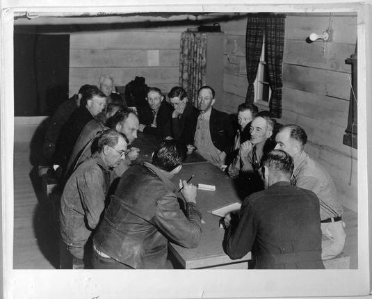 The image depicts a historical photograph of fifteen men seated around a table in what appears to be an indoor setting, likely from the early 20th century given its black and white composition. The room has wooden paneling on the walls with curtains hanging at one side.

Most of the individuals are dressed in dark-colored clothing such as jackets or coats; some wear glasses. A few men have their hands clasped together while others appear to be looking down, possibly reading documents or writing notes. At least two men hold pens and a notebook is visible on the table.

The setting suggests an organized meeting or discussion taking place among these individuals who may represent different roles within this group. The overall atmosphere of the image conveys seriousness and engagement with their topic at hand.