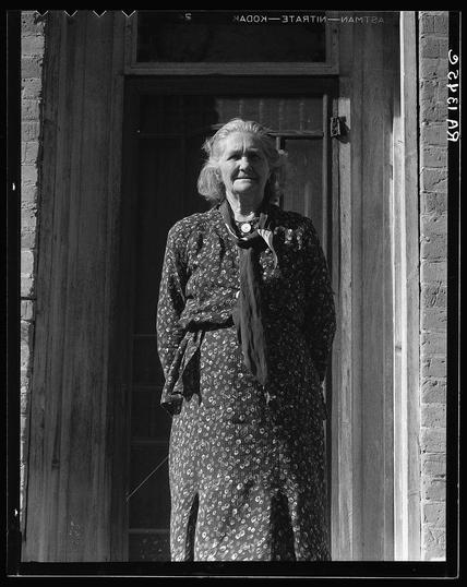 The image depicts a middle-aged woman standing in the doorway of what appears to be an older, wooden structure. She is dressed in period attire that suggests early 20th-century fashion: she wears a long-sleeved floral print dress with dots and dark fabric belt cinching at the waist. Over her shoulders, she has draped a shawl or scarf adorned with round patterns. Her hair is parted on one side, and it falls over what looks like a white collar beneath the dress's neckline.

The photograph itself appears to be in black and white, which adds a sense of historical authenticity to the image. The quality of light suggests that this photo was taken outdoors during daylight hours; however, specific environmental conditions or time are not discernible from the provided details. There is text at the top left corner on what seems like a title card reading "Kodak - State Art Museum," followed by some illegible script and numbers.

This image evokes historical context related to early education in rural America during the late 19th or early 20th centuries, as indicated by the caption mentioning her being a schoolteacher.
