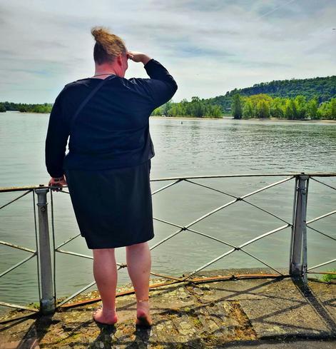 Rear view — A barefoot person standing at the very edge of an old stone platform by the Rhine river in Rüdesheim, wearing a simple black skirt and a matching black top. They lean slightly forward against a weathered metal railing, one hand shielding their eyes from the soft midday sun as they gaze thoughtfully across the wide, glistening river. Their tousled hair is pulled up into a playful, fluffy ponytail, and a delicate anklet encircles their left ankle, catching the light. Their bare feet are firmly grounded on the rough stone ... the left foot flat, while the right foot is raised gracefully onto its toes, creating a subtle, dynamic tension that mirrors the soft movement of the breeze. The horizon reveals lush green trees and a low, forested hill bathed in spring's fresh colors.