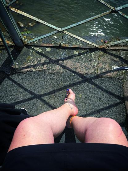 Fist-person view — Looking down over sun-kissed bare legs stretched out carefreely on the warm, rough pavement next to the river. The camera captures the crossed ankles perfectly, one foot slightly raised, the toes painted in a bold, playful blue. An elegant silver anklet with tiny charms decorates the left ankle, while a vibrant orange ankle band above the silver band adds a cheeky splash of color on the right. The ground is a tapestry of old asphalt and worn stones, framed by the rusty, crisscrossing bars of the metal railing, their shadows dancing lazily across the legs and ground.
To the left a shoe can be spotted.