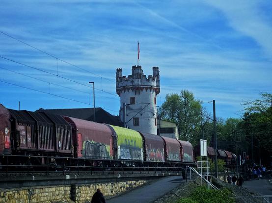 Street-level view — A freight train, adorned with colorful graffiti, rumbles past a whimsical, medieval-style white tower in Rüdesheim, its battlemented crown jutting confidently against a vivid, lightly clouded blue sky. The train’s muted reds and a single, strikingly neon-green container create a raw, urban contrast to the historical setting. In the foreground, people stroll casually along a stone-lined riverside path, some caught mid-step, enjoying the breezy day. Overhead, delicate lines of the railway's catenary system carve soft arcs into the expansive sky.