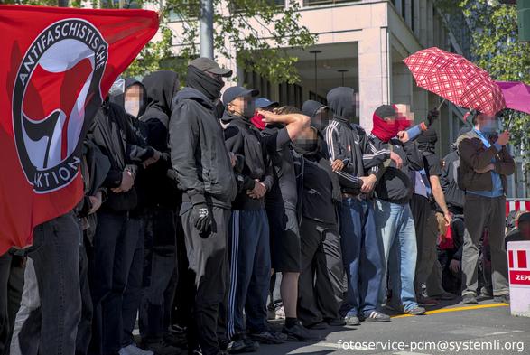 boacke mit menschen die zum teil vermummt und untergehakt auf der straße stehen,