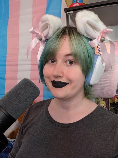 Selfie of Revy wearing black lipstick and purple and white headphones with white pink fluffy cat ears attached 
She's near a microphone