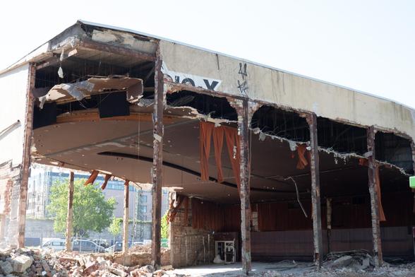 The auditorium of an old cinema that is currently being demolished. Three of the outer walls have already been removed, leaving only steel girders to support the roof. The auditorium is empty and long, brown shreds of wall panelling hang from the ceiling. They move in the wind