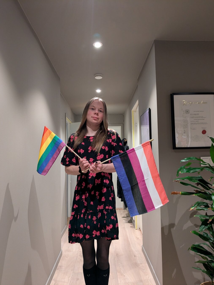 A woman stands in her apartment.

She is holding a LGBTQ+ flag and a genderfluid flag. 

In the foreground, the woman stands in a hallway with an indoor plant visible to the right and a certificate on the wall behind.