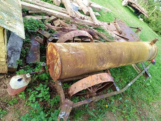 A long, cylindrical steel tank on top of a steel wheeled trailer. Under the spout with open valve, a small plastic jerrycan about half full with a yellow/brown fluid. On the far end is a filler/vent cap. There's some other junk around.
