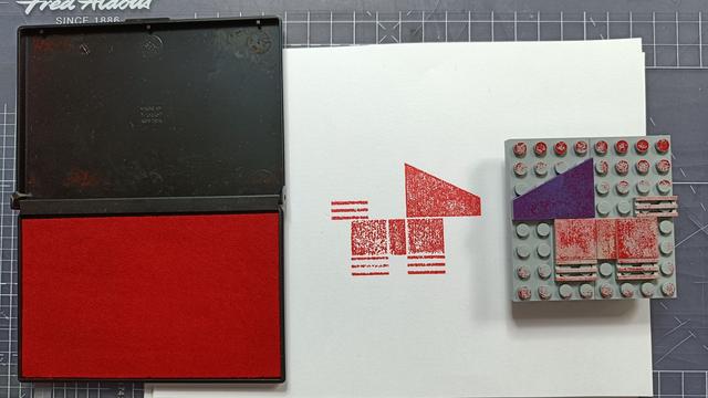 On the left a red inkpad, on the right a Lego design using flat plates that is a dog facing left. In the middle on a sheet of white paper a red dog has been printed, facing right.