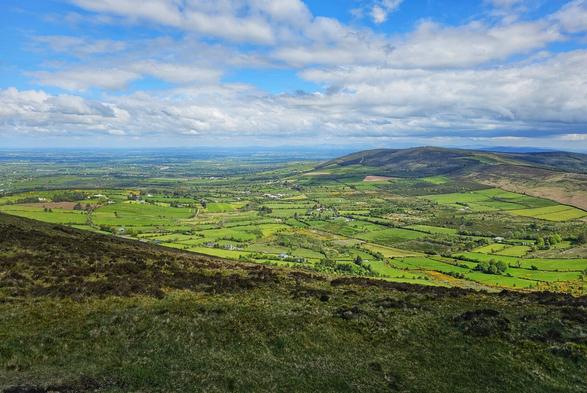 A wide shot from a high vantage point shows a vast expanse of green rolling hills and farmland under a partly cloudy blue sky.