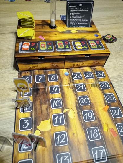Die ausgeklappte Verpackung offenbart ein nummeriertes Spielfeld sowie einen Aufbau mit zwei Schubladen, in denen sich die Fragekarten sowie Meeples etc. befinden.