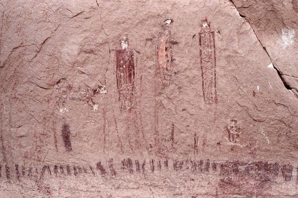 A dog and three thin anthropomporphic figures painted in red on a sandstone wall.
