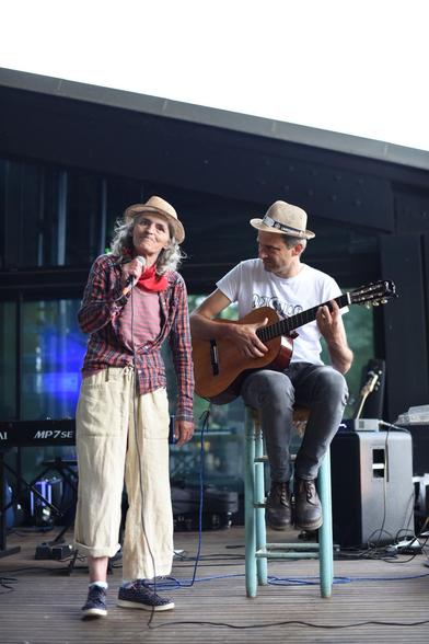 Scène en extérieur. Une femme debout tient un micro, elle porte un chapeau de paille. À droite un homme avec un autre chapeau de paille est assis sur un tabouret haut, une guitare classique dans les bras.
