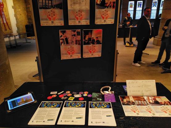 People standing around a pin up wall with photos and information about hackathons, a tablet, stickers, name badges, and short overview of the process.