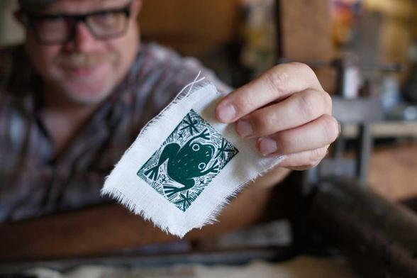 ken leaning over a press. holding a piece of fabric with a smiling frog on it