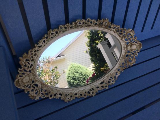 An oval old-fashioned white-ish metal ornate frame or vanity tray has had its center replaced with a mirror.  It is sitting propped up (horizontally) on a light blue polywood bench in the shade.  

The mirror itself is reflecting a bright scene of yellow house, green crepe myrtle and some kind of closely-trimmed conical evergreen.  It looks a little like a small porthole to another, brighter world.

This date.  Eight years ago.