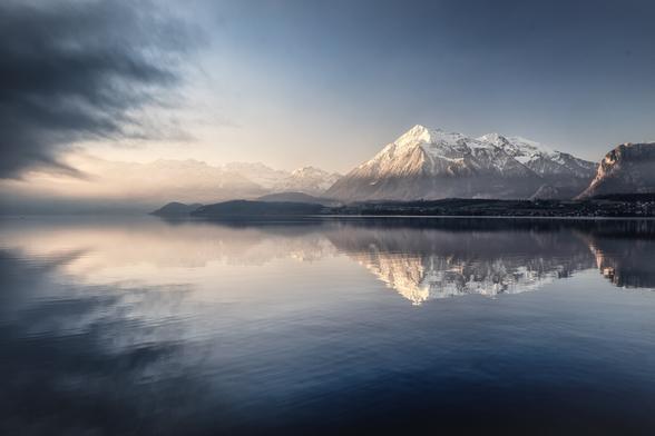 Thunersee mit Niesen, der sich auf der Wasseroberfläche spiegelt. Vom linken Rand greift Nebel ins Bild.