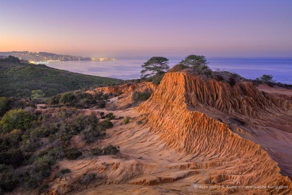 A color landscape photo showing an eroded sandstone formation in soft pre-dawn light. Beyond it, the sweeping view follows a curved coastline to distant city lights.
