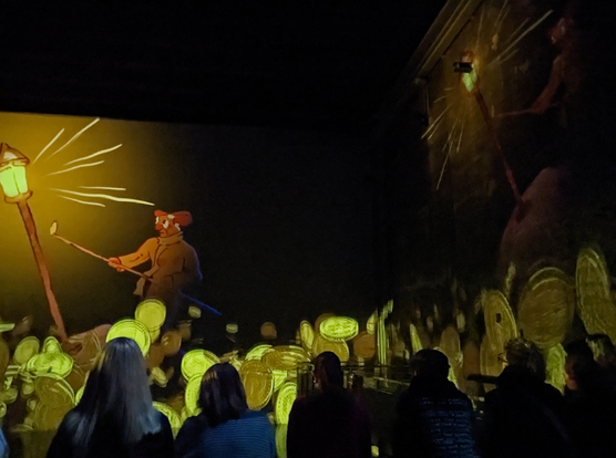 the motion picture of a man lighting a candle and gold coins underneath projected on two walls while bystanders, real spectators of the show (Le petit prince), watch. 

Nicolas Mouart, all rights, 2024