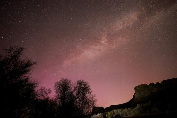 A nightscape of part of the Milky Way band and surrounding stars, washing over into a unscheduled purple/magenta sky. In the distance is the glow of the bright lights of Coulee City, Washington. On the right, you can make out the faint outlines of a dramatic butte in the distance. On the left are the dark outlines of some trees in the foreground. To the right of them, there's sagebrush.
