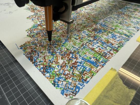 The image shows a pen plotter machine in the process of creating a detailed, colourful geometric pattern on paper. The pattern consists of interlocking, maze-like lines in blue, green, brown, and red. The paper is secured with yellow tape on a gridded cutting mat.
