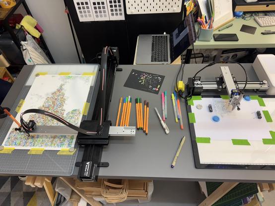 The image shows a workspace with two pen plotter machines. On the left, one plotter is working on a colourful design. On the right, another plotter is set up with blank paper, markers, and pens ready for use. A laptop is partially visible behind the machines.
