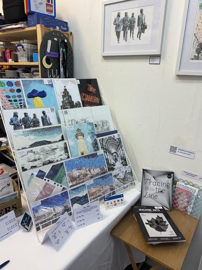 A display at an art or craft fair stall. Various art prints and postcards are arranged on a large board, showcasing diverse designs, including landscapes, architectural elements, and abstract illustrations. Some images feature notable landmarks, such as lighthouses and cityscapes, while others have stylised portraits and creative patterns.