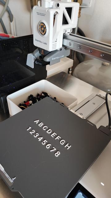 View on a A1Mini 3D filament printer showing small white letters and numbers printed on a Bambulab SuperTack buildplate. The numbers 1 through 8 and letters A through H for a chessboard project.