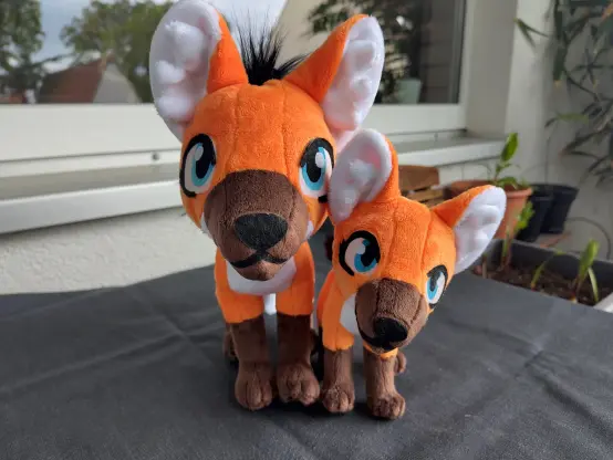 Two different sized maned wolf plushies made from orange, white and brown fabric next to each other. The smaller one is roughly half the size.