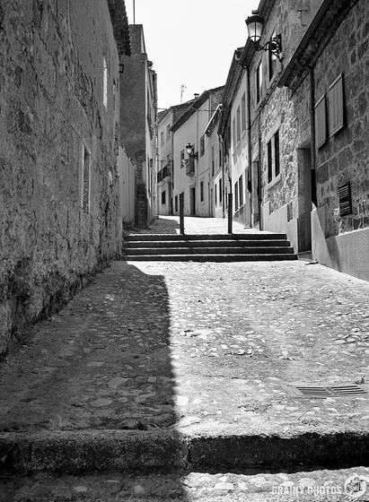 A black and white photo of a narrow, uphill cobblestone street lined with old buildings. Lamps mounted on the walls along the empty street, extend into the distance and evoke a sense of historical charm.