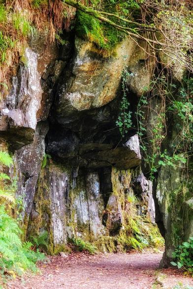 A rocky path leads through a natural archway formed by large moss-covered rocks, with ferns and other greenery visible.