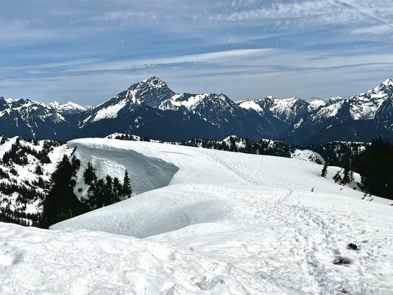 View of more peaks from the top of Dickerman. The foreground shows a snowy flat area and a snow cornice-- where the crusty snow forms a diving board like extension out over a sheer cliff.