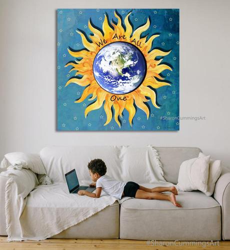 A sun symbol with an earth overlay on a night sky with stars.  It reads "We Are All One".  In a furniture setting with a little child on a laptop on a sofa.  Art by Sharon Cummings.