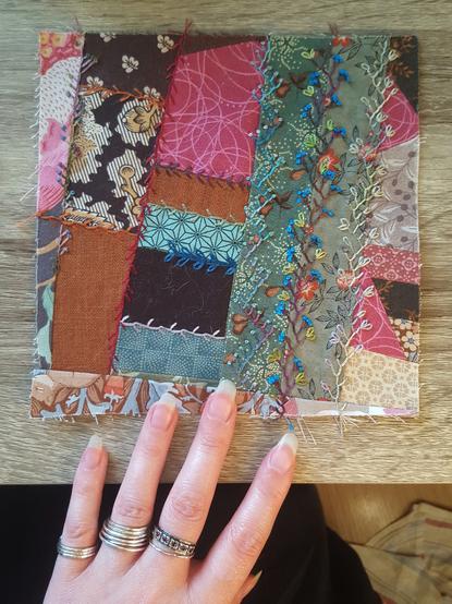 Top down photo of an embroidered quilt block, comprised of chaotically different fabric scraps, of almost all possible colours but earth and plant tones mostly. There's different stitches making up little vines, plants and flowers trailing along seams, less and more complex. There's a couple of subtly variegated threads and a couple of brighter single tone ones, most noticeably a deep lapis lazuli blue, almost electric blue on its backdrop of green toned botanical prints. It's pretty eclectic all in all, but somehow pulls together in its chaos. Sini's pale hand in frame, with her signature silver rings.