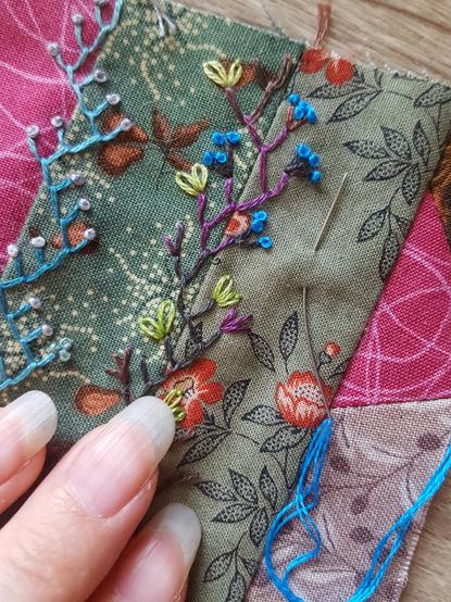 A very close up photo of the little trailing vine with the electric blue thread. It's like a pretty abstracted take on a blueberry plant, with brown and purple variegated thread in feather stitch for the stem and twigs, a very vibrant light spring green for the leaves in lazy daisy stitch, and the very very vibrant blue little berries in French knots. It's still in progress, with sewing needle and its threaded blue embroidery floss, just exceptionally blue and standing out against the other more muted tones. It's a very pretty colour. The fabrics are green with red roses and ripe golden pears on them, flanked by bright pink. It sounds wilder than it looks! Sini's fingertips in frame, with slightly overlong oval natural nails.
